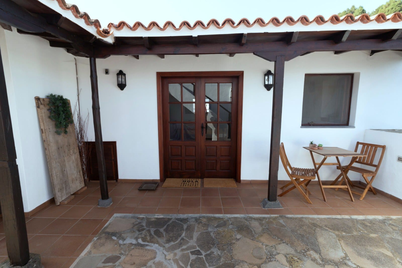 Entrada principal de Finca Julia bajo la pérgola de madera, con puerta acristalada, felpudo y una mesa para dos en el patio de losa