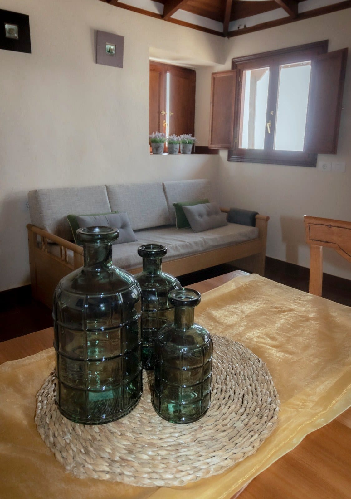 Sala de estar de Finca Julia con banco de madera y cojines, ventana con contraventanas de madera y botellas de vidrio verde sobre la mesa